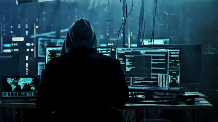 imagem de um hacker em frente a um computador com uma tela cheia de códigos, representando fraudes digitais e cibercriminosos