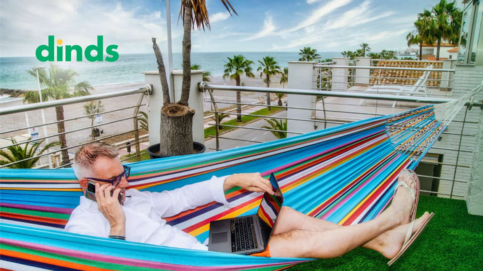 homem fala ao celular com notebook no colo sentado em uma rede em frente a um praia