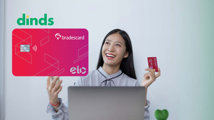mulher sorri segurando cartão de crédito e celular com notebook aberto e cartão bradesco elo internacional ao lado