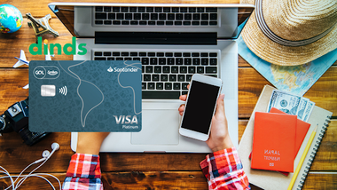 pessoa com celular e computador sentado em uma mesa, com caderneta, dólares, e passaporte, o cartão de crédito gol smiles platinum está ao lado, logo abaixo do logo Dinds