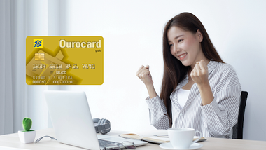 mulher em seu escritorio com uma xícara de café, com seu computador comemorando o cartão de crédito ourocard visa gold