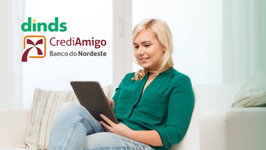 mulher sentada no sofá, usando tablet ao lado dos logos do Dinds e CrediAmigo Banco do Nordeste