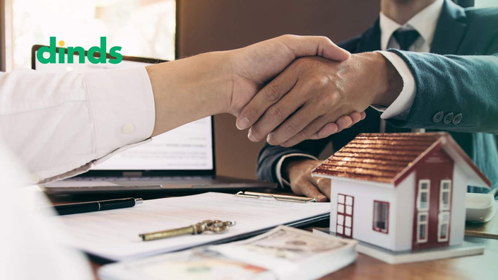 duas maõs se apertam com maquete de casa e dinheiro e chave e papel em cime ada mesa