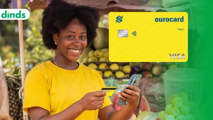 mulher sorri segurando cartão e celular nas mãos em frente a barraca de frutas com cartão ourocard fácil ao lado