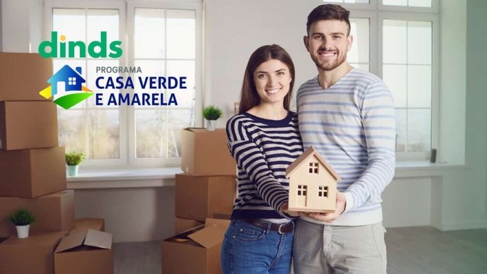 Casal segurando casa de madeira com programa casa verde e amarela ao lado e caixas de papelão ao lado