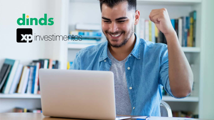 homem sorri e comemora olhando para tela de notebook com estantes de livros atrás