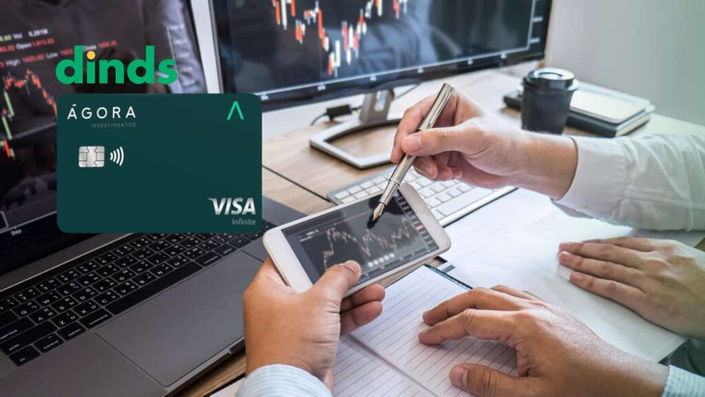 Dois investidores analisando gráficos financeiros em uma tela e dispositivo móvel, com imagem do cartão Ágora Investimentos Visa Infinite e logo do Dinds.