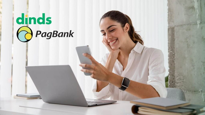 Mulher sorrindo com celular na mão e notebook numa mesa