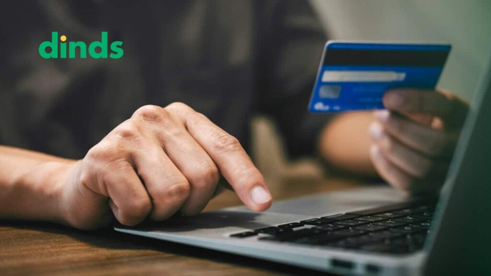 Mão segurando cartão de crédito enquanto realiza uma compra online em um laptop, com logo do Dinds.