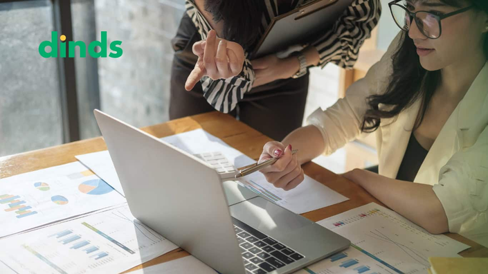 duas mulheres apontam para tela de notebook com papéis com gráficos na mesa