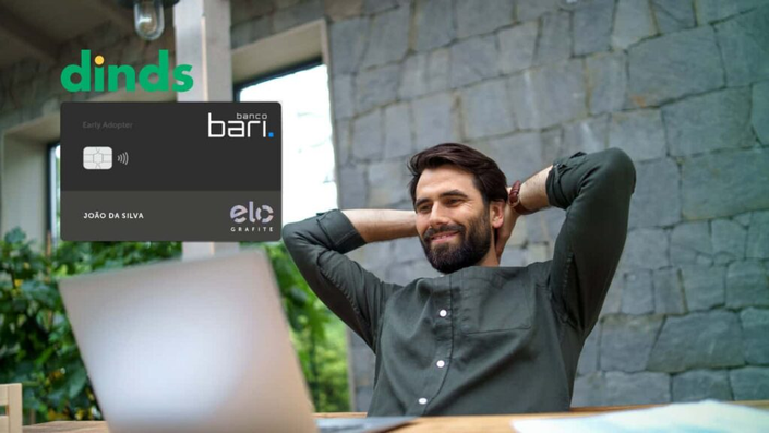 Homem sorridente relaxando em frente ao laptop, ao lado da imagem do cartão Banco Bari Elo Grafite e logo do Dinds.